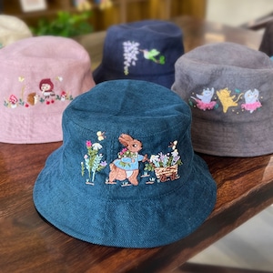 May include: A collection of six embroidered bucket hats in various colors, including pink, blue, gray, and green. Each hat features a different embroidered design, including a rabbit, a squirrel, and a bird.