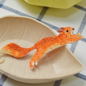May include: A small, orange embroidered squirrel applique. The squirrel is in a jumping pose with its tail curled behind it.