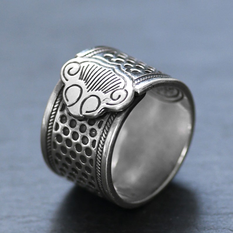 Puede incluir: Un dedal de plata con un dise&ntilde;o decorativo en la parte superior. El dedal tiene una superficie texturizada y una forma redonda.