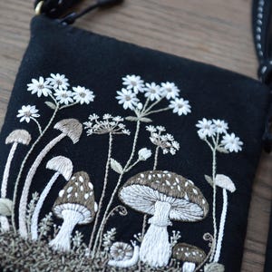 May include: A black crossbody bag featuring detailed embroidery of various mushrooms, small white flowers, and a snail. The design uses white, beige, and brown thread on a dark fabric base. The bag includes a black strap with white decorative stitching.