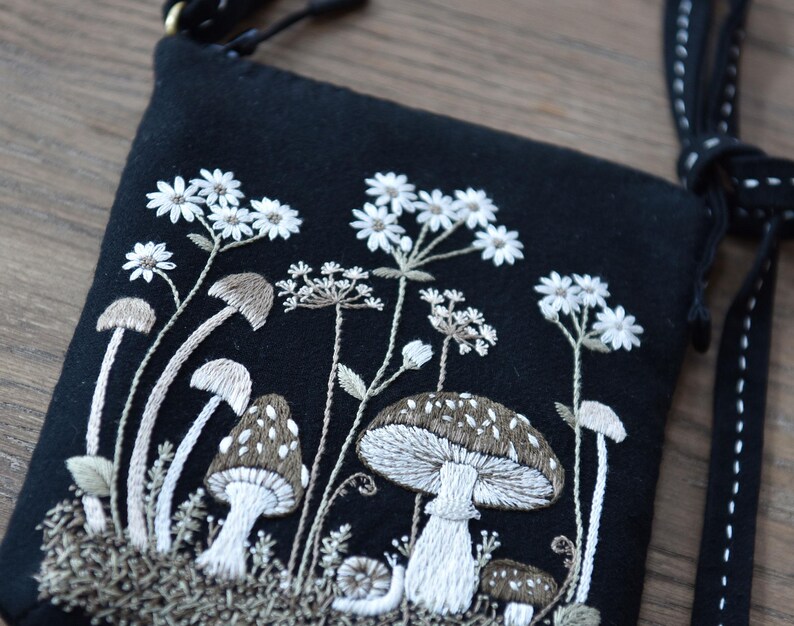 May include: A black crossbody bag featuring detailed embroidery of various mushrooms, small white flowers, and a snail. The design uses white, beige, and brown thread on a dark fabric base. The bag includes a black strap with white decorative stitching.