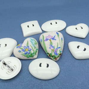 Op de afbeelding: Assortiment van witte keramische broches met bloemenborduurwerk. De broches zijn in de vorm van harten, ovalen en vierkanten. Het borduurwerk bevat blauwe, paarse en groene bloemen.