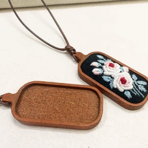 Pu&ograve; includere: Due collane con ciondoli in legno. Un ciondolo presenta uno sfondo nero con rose bianche e rosse ricamate e foglie blu. L'altro &egrave; un rettangolo di legno liscio. Entrambi sono attaccati a cordini marroni.