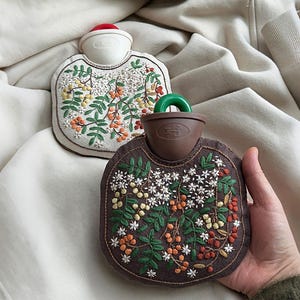 Może przedstawiać: Dwa haftowane termofory. Jeden jest biały z czerwonym korkiem, drugi brązowy z zielonym uchwytem. Oba mają kwiatowe hafty w kolorze zielonym, pomarańczowym i białym. Termofory leżą na kremowej tkaninie.