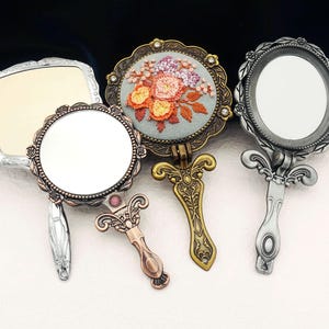 May include: Three ornate hand mirrors with different designs. One mirror has a floral embroidery design, another has a silver frame with a leaf design, and the third has a plain silver frame.