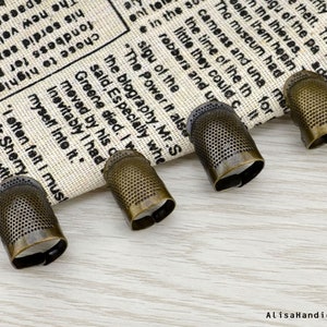 May include: Four antique gold-toned thimbles with a textured surface and a circular opening. The thimbles are arranged in a row on a white fabric background.