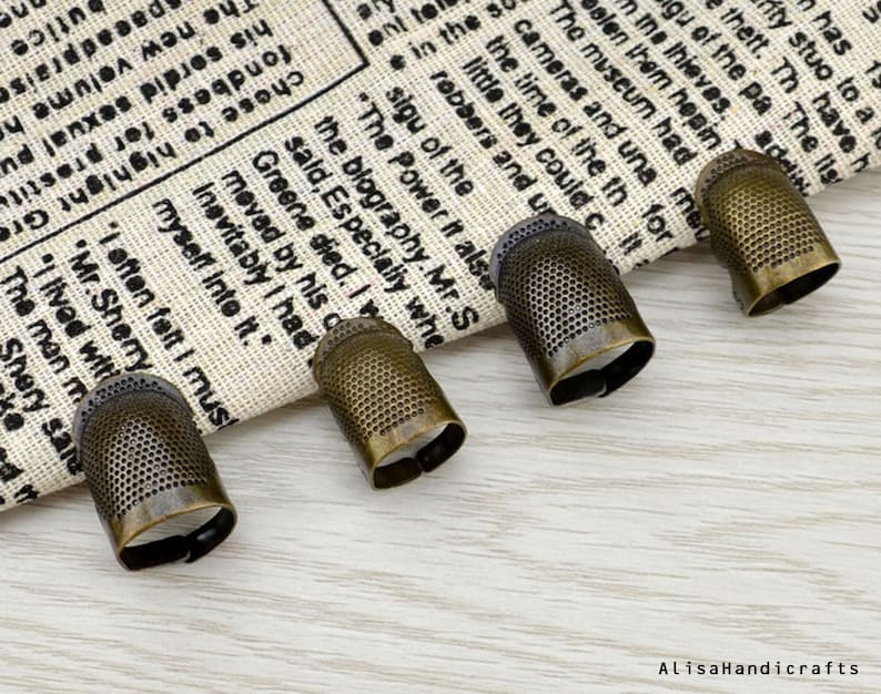 May include: Four antique gold-toned thimbles with a textured surface and a circular opening. The thimbles are arranged in a row on a white fabric background.