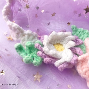 May include: A close-up of a crocheted flower crown with white, pink, light green, and purple flowers. The crown is on a purple tulle background with gold glitter stars.