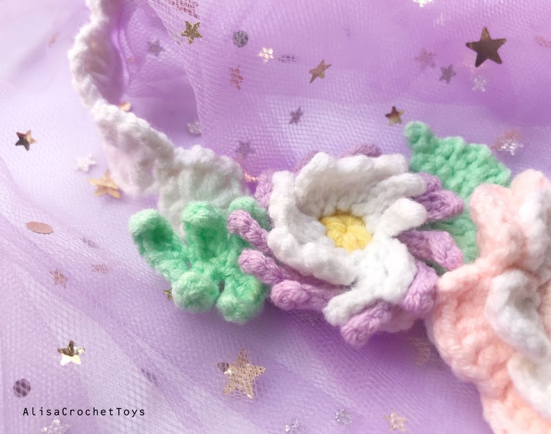 May include: A close-up of a crocheted flower crown with white, pink, light green, and purple flowers. The crown is on a purple tulle background with gold glitter stars.