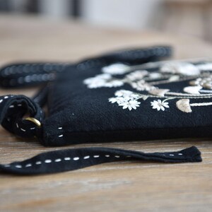 May include: A black crossbody bag featuring white and beige floral embroidery on the front. The bag has a long black strap with white running stitch detailing and a brass-colored metal ring attachment on the side of the bag structure.