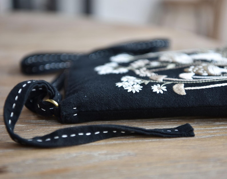 May include: A black crossbody bag featuring white and beige floral embroidery on the front. The bag has a long black strap with white running stitch detailing and a brass-colored metal ring attachment on the side of the bag structure.