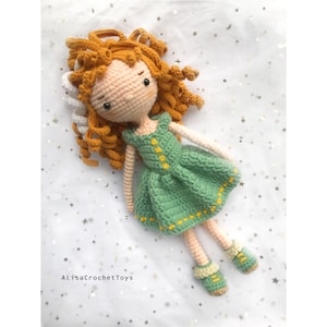 May include: A crocheted doll with curly orange hair, wearing a green dress with yellow accents and white booties. The doll is lying on a white background with gold stars.