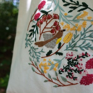 Puede incluir: Una bolsa de tela blanca con un dise&ntilde;o bordado que presenta un p&aacute;jaro, flores y hojas en tonos de verde, amarillo, rojo y marr&oacute;n.