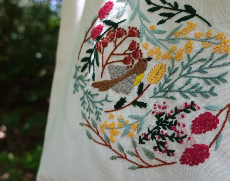 Puede incluir: Una bolsa de tela blanca con un dise&ntilde;o bordado que presenta un p&aacute;jaro, flores y hojas en tonos de verde, amarillo, rojo y marr&oacute;n.