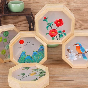 May include: Four octagonal wooden frames with embroidered designs. The frames are on a wooden surface. The designs include a bird, flowers, mountains, and a plant.