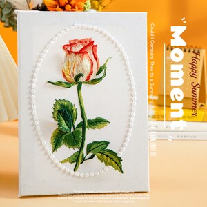 Puede incluir: Un lienzo blanco con una sola rosa bordada en tonos rojos, naranjas y amarillos. La rosa está rodeada por un borde de perlas. El texto "Could I Compare Thee to a Summer's Day." está impreso en el lado derecho del lienzo. El texto "Moment" está impreso verticalmente en letras grandes en el lado derecho del lienzo. El texto "Happy Summer" está impreso horizontalmente sobre la palabra "Moment".