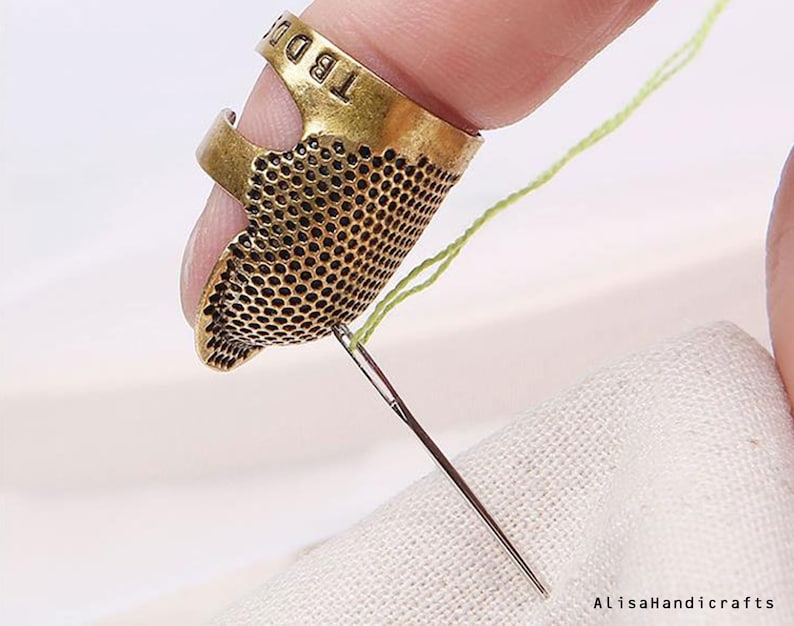 May include: A gold metal thimble with a textured surface and a ring to hold it on a finger. The thimble is being used to push a needle through a piece of white fabric.