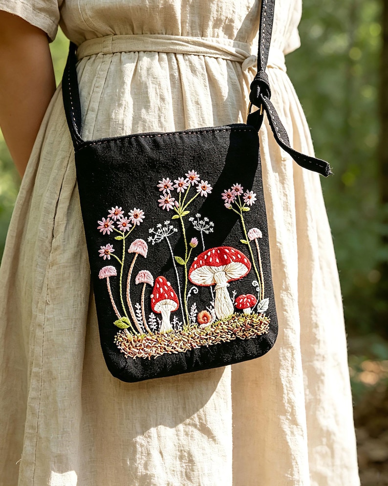 May include: A black crossbody bag featuring detailed embroidery of red and white mushrooms, small pink flowers, and green foliage. The bag has a long black strap and is worn over a light beige dress. The embroidery covers the front of the bag.