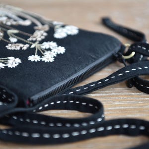 May include: A black fabric crossbody bag featuring white and beige floral embroidery on the front. The bag has a black zipper closure and a long black strap decorated with a white dashed stitch pattern running along the entire length of the strap.