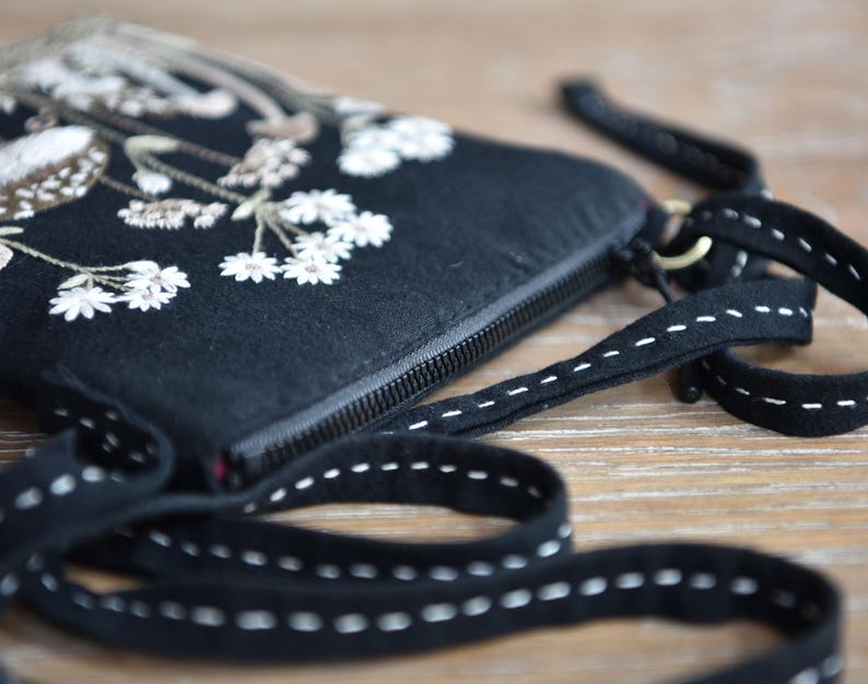 May include: A black fabric crossbody bag featuring white and beige floral embroidery on the front. The bag has a black zipper closure and a long black strap decorated with a white dashed stitch pattern running along the entire length of the strap.
