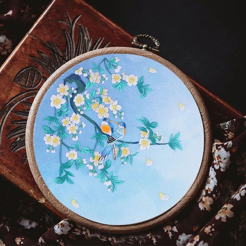 May include: Embroidered hoop art featuring two blue birds perched on a branch with white blossoms. The background is a light blue gradient with scattered white petals.
