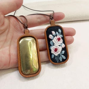 Puede incluir: Dos colgantes rectangulares con marcos y cordones de cuero marrón. Uno tiene un centro dorado pulido. El otro tiene un fondo negro con flores rosas y blancas bordadas y hojas azules.