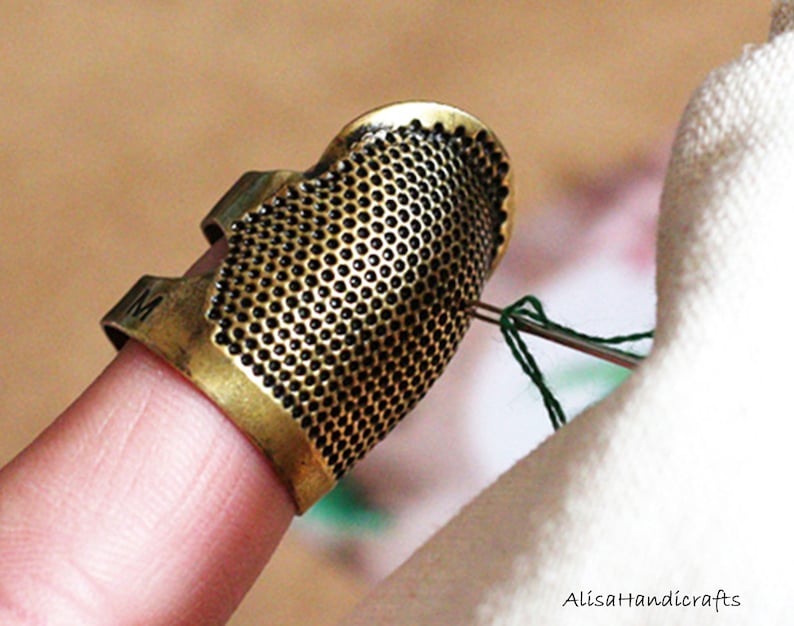 May include: A gold metal thimble with a textured surface and a small opening on the side. The thimble is worn on a finger and is being used to thread a green needle.