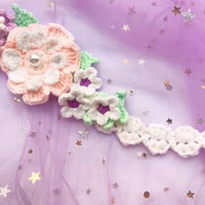 May include: A white crocheted flower crown with pink and purple flowers and green leaves. The crown is on a purple background with silver glitter stars.