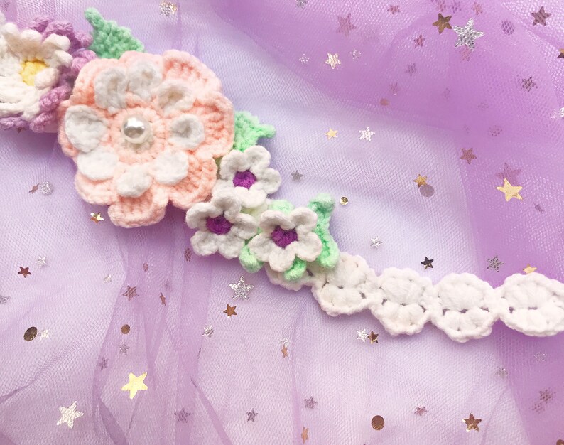 May include: A white crocheted flower crown with pink and purple flowers and green leaves. The crown is on a purple background with silver glitter stars.