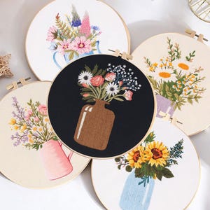 May include: A collection of five embroidered floral designs in wooden hoops. The designs feature various flowers in vases and pitchers, with a color palette of pink, yellow, blue, and white. The hoops are displayed on a white surface.