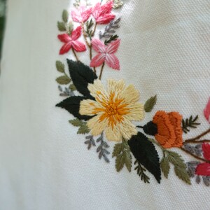Puede incluir: Dise&ntilde;o floral bordado sobre un fondo de tela blanca. El dise&ntilde;o presenta flores amarillas, naranjas, rosas y verdes.