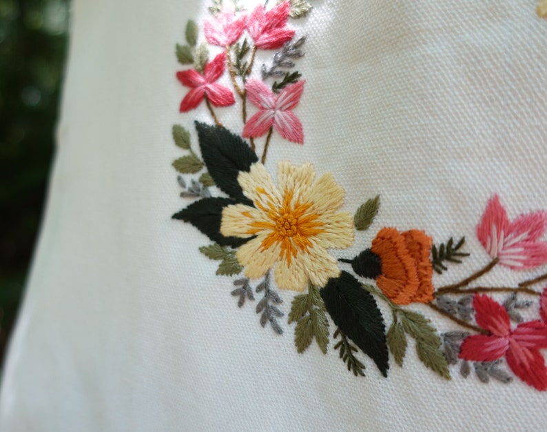 Puede incluir: Dise&ntilde;o floral bordado sobre un fondo de tela blanca. El dise&ntilde;o presenta flores amarillas, naranjas, rosas y verdes.