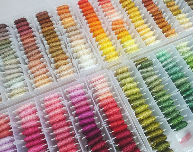 May include: A close-up of a collection of embroidery floss spools organized in a clear plastic storage box. The spools are arranged in rows by color, with shades of brown, orange, yellow, green, pink, and purple.