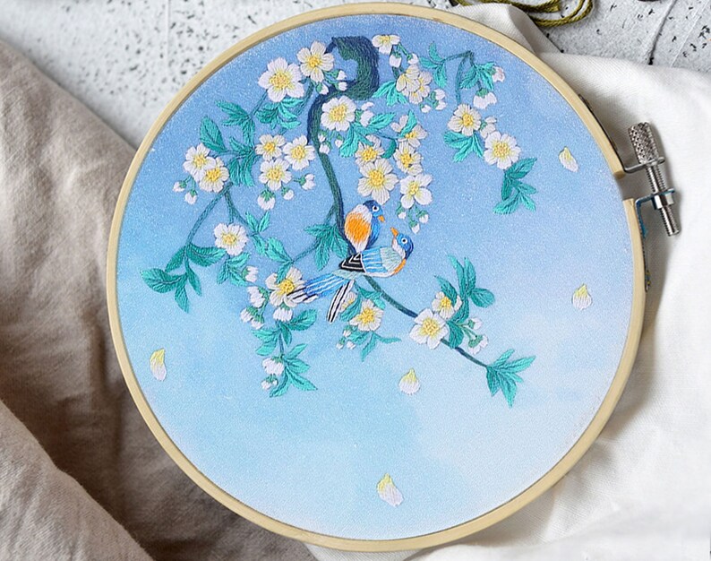 May include: Embroidered hoop art featuring a blue sky with white blossoms and two blue birds on a branch.