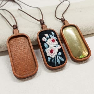 Pu&ograve; includere: Tre collane con ciondoli in legno con cordini marroni. Un ciondolo &egrave; vuoto, uno ha un inserto color oro e l'altro presenta uno sfondo nero con fiori bianchi e rosa ricamati e foglie blu. I ciondoli sono rettangolari con bordi arrotondati.