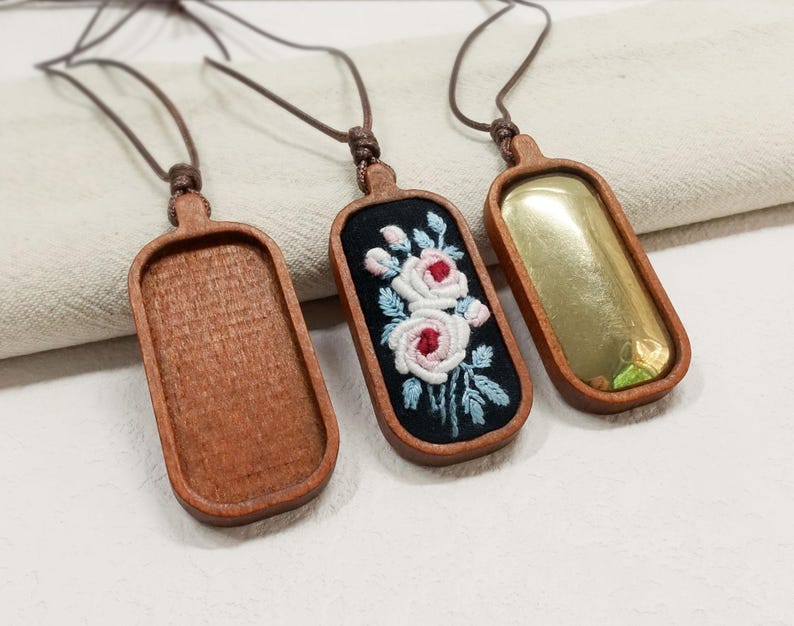 Pu&ograve; includere: Tre collane con ciondoli in legno con cordini marroni. Un ciondolo &egrave; vuoto, uno ha un inserto color oro e l'altro presenta uno sfondo nero con fiori bianchi e rosa ricamati e foglie blu. I ciondoli sono rettangolari con bordi arrotondati.