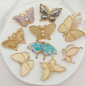 Op de afbeelding: Een verzameling vlinderbroches in goud, zilver, paars en blauw. Sommige zijn versierd met strass-steentjes en parels. De broches zijn in verschillende ontwerpen en worden gepresenteerd op een witte ondergrond.