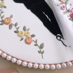 Pu&ograve; includere: Un cerchio da ricamo bianco con un bordo di perle, che presenta un delicato disegno floreale ricamato in tonalit&agrave; di rosa, giallo e verde. Il disegno include piccole rose e foglie.