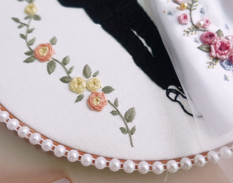 Pu&ograve; includere: Un cerchio da ricamo bianco con un bordo di perle, che presenta un delicato disegno floreale ricamato in tonalit&agrave; di rosa, giallo e verde. Il disegno include piccole rose e foglie.