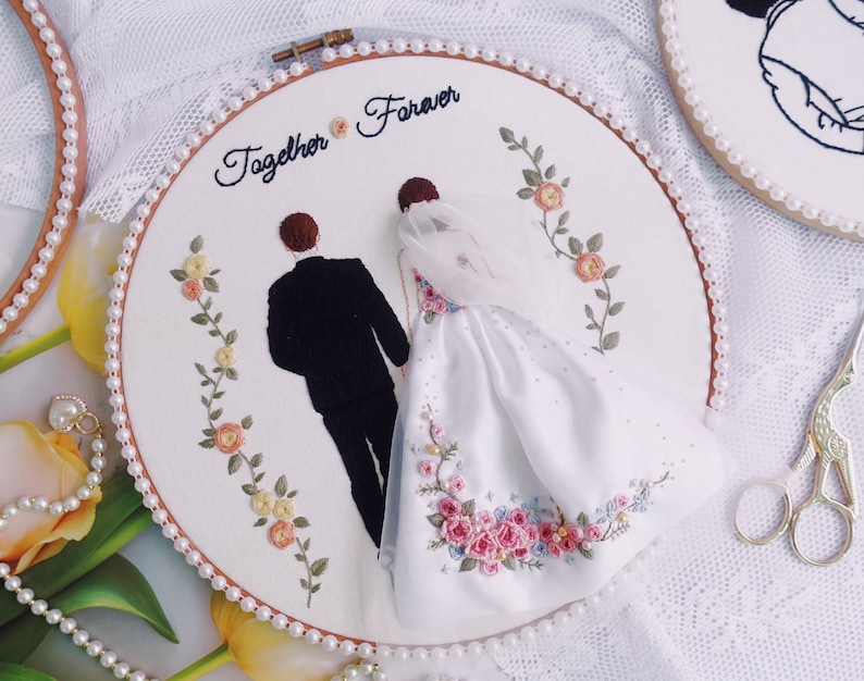 Pu&ograve; includere: Arte di ricamo su telaio con sfondo bianco e bordatura di perle. Il telaio presenta una coppia, uno sposo in abito nero e una sposa in abito bianco, con il testo "Together Forever" sopra di loro.