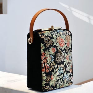 May include: A black handbag with a floral tapestry design. It features a brown leather handle and gold-coloured hardware. The floral pattern includes red, green, and blue flowers. The bag is rectangular in shape.