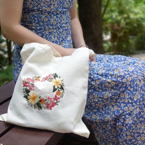 Puede incluir: Una bolsa de tela blanca con un dise&ntilde;o de bordado floral en forma de coraz&oacute;n. La bolsa est&aacute; hecha de un tejido suave y natural y tiene un dise&ntilde;o simple y elegante.