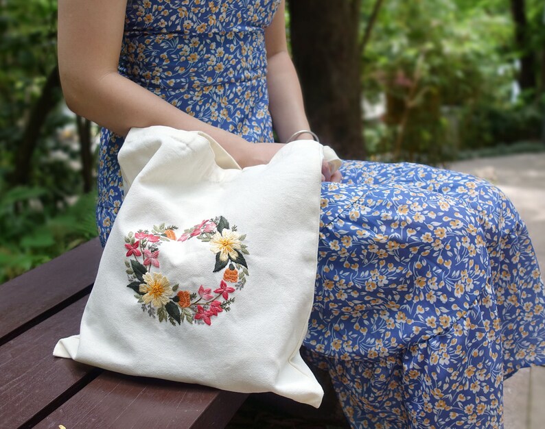 Puede incluir: Una bolsa de tela blanca con un dise&ntilde;o de bordado floral en forma de coraz&oacute;n. La bolsa est&aacute; hecha de un tejido suave y natural y tiene un dise&ntilde;o simple y elegante.