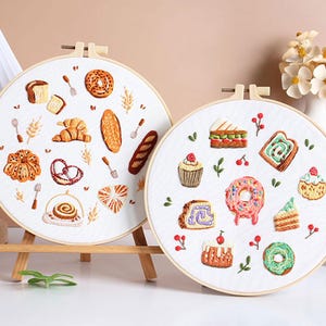May include: Two circular embroidery hoops displaying hand-stitched images of baked goods. One shows bread, a croissant, and a pretzel. The other features a donut, cupcakes, and cake slices. Both are set on wooden stands.