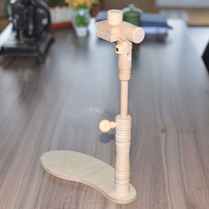 May include: A wooden embroidery stand with a base and a vertical post that adjusts in height. The stand has a knob for tightening the post and a clamp at the top for holding embroidery hoops.