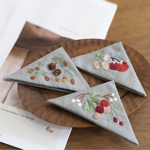 Puede incluir: Tres piezas triangulares de tela con diferentes diseños bordados. La primera pieza tiene un diseño marrón, verde y amarillo de piñas y bellotas. La segunda pieza tiene un diseño rojo, verde y blanco de cerezas y hojas. La tercera pieza tiene un diseño rojo, verde y blanco de cerezas y hojas.