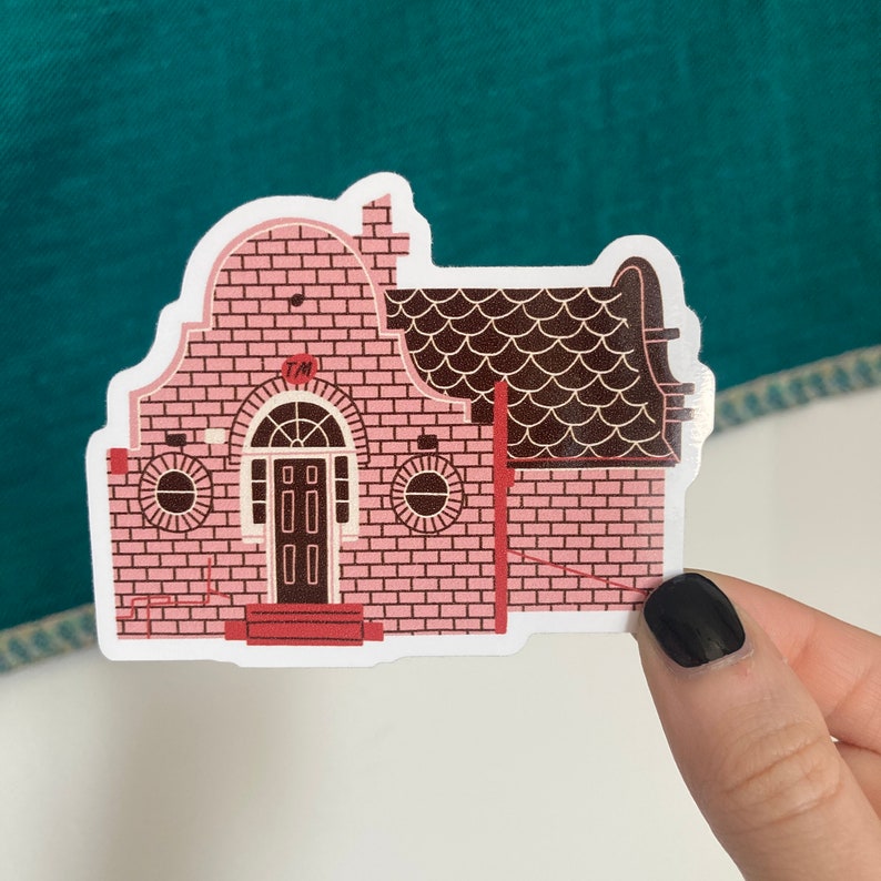 May include: A sticker depicting a pink brick house with a brown roof and door. The house has two circular windows and a red doorstep. The sticker has a white border and is held by a hand.