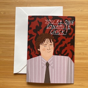 May include: Greeting card featuring a cartoon illustration of a man in a striped shirt and tie. The card has the text "YOU'RE ONE DYNAMITE CHICK!" on a red and black patterned background. A white envelope is behind the card.