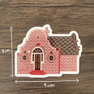 May include: A sticker of a pink brick house with a brown roof and door. The sticker is 9 cm wide and 8 cm tall. The house has a chimney, round windows, and a small porch. The sticker has a white border.