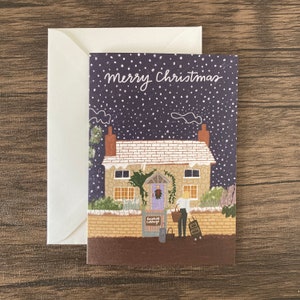 May include: A Christmas card with a cottage illustration. The card features a snowy scene with the text "Merry Christmas" in white script. A white envelope is next to the card. The cottage has a purple door and the name "Rosehill Cottage" on the gate.